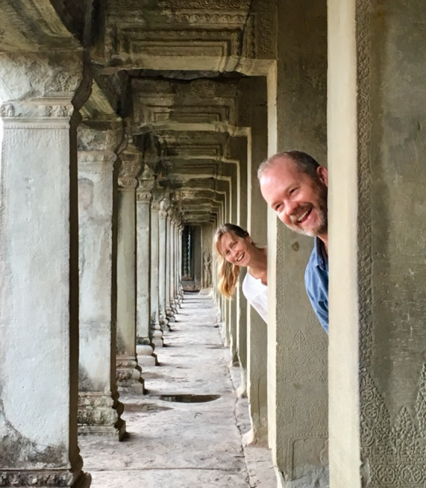In a long hallway of columns, a man and woman playfully peek out from behind the architecture.