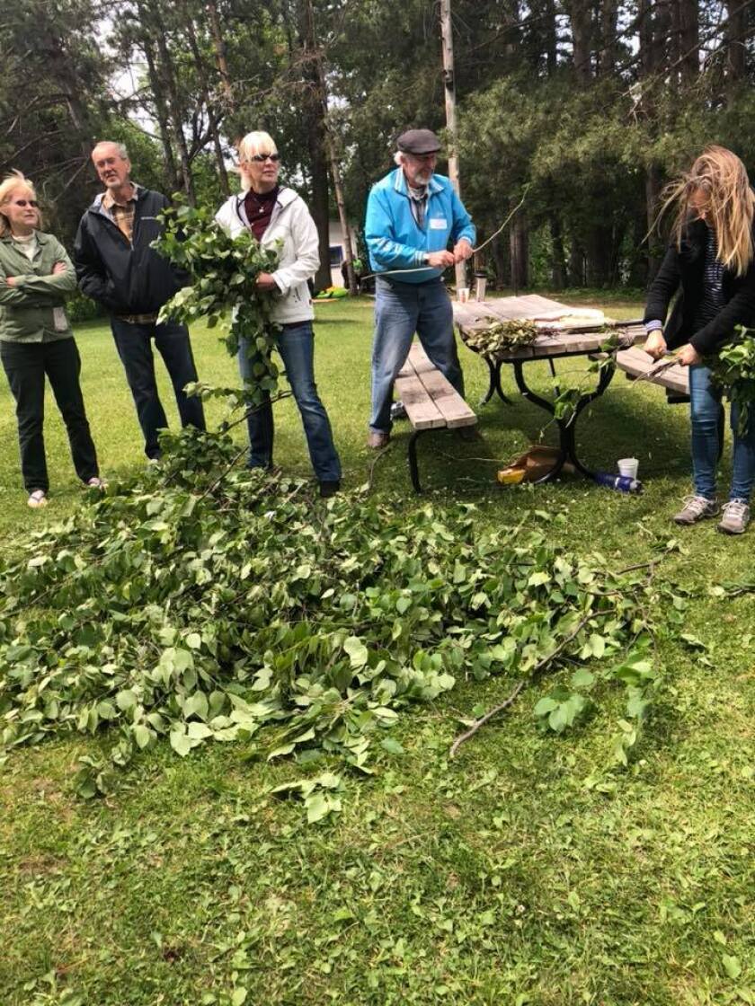 Five people stand outdoors over a pile of gathered tree branches, trimming and lashing them into clusters.