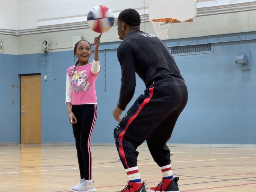 Flip White, wearing Harlem Globetrotters warmup suit, leans forward as he helps a smiling young girl spin a basketball on her finger.