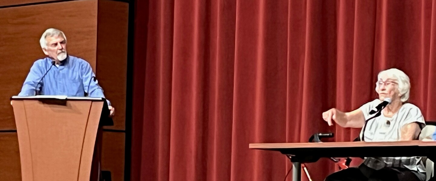 Dave Johnson stands at a podium on stage at the CTC Center in Pillager on Thursday, July 28, 2022, while his aunt and retired rural school teacher Ruth Johnson Boldan sits at a nearby table during a panel discussion about education.