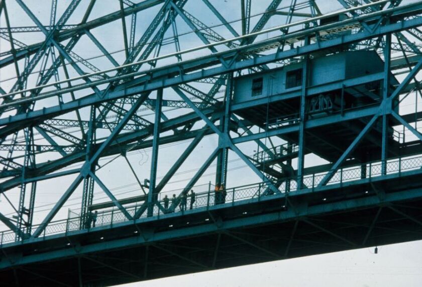 View from below of Aerial Lift Bridge lower span, with a handful of riders visible in a chain-link cage along the pedestrian walkway.