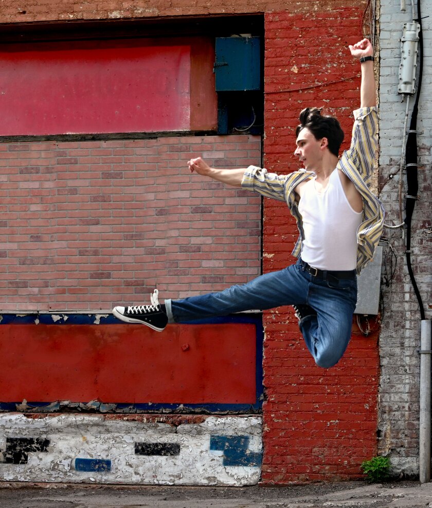 A man wearing blue jeans, black Converse All-Star sneakers, a white t-shirt, and an open striped button-down shirt dances energetically against a red-painted brick wall.