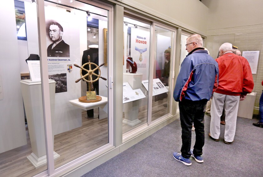 An elderly white man stands looking at a museum display containing artifacts including the small steering wheel of a ship.