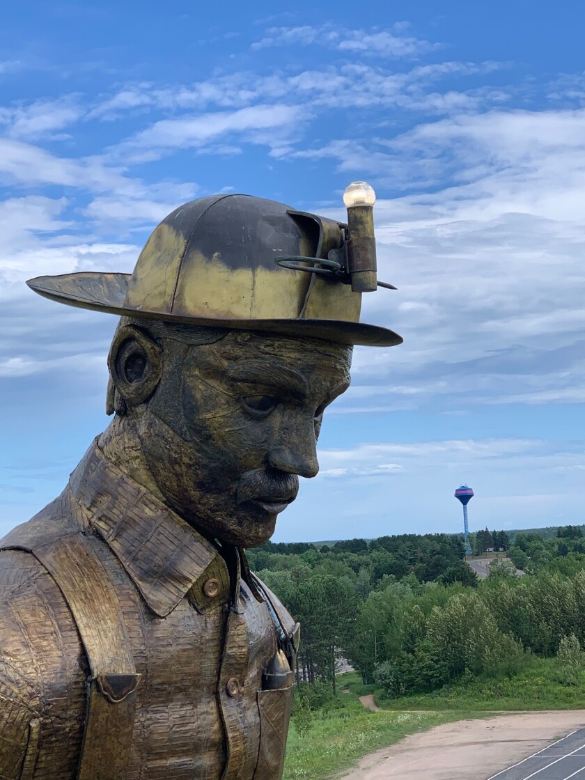 Face of a large statue of an iron ore miner, with a helmet lamp illuminated. Warm summer weather is in evidence, with green trees and blue skies.