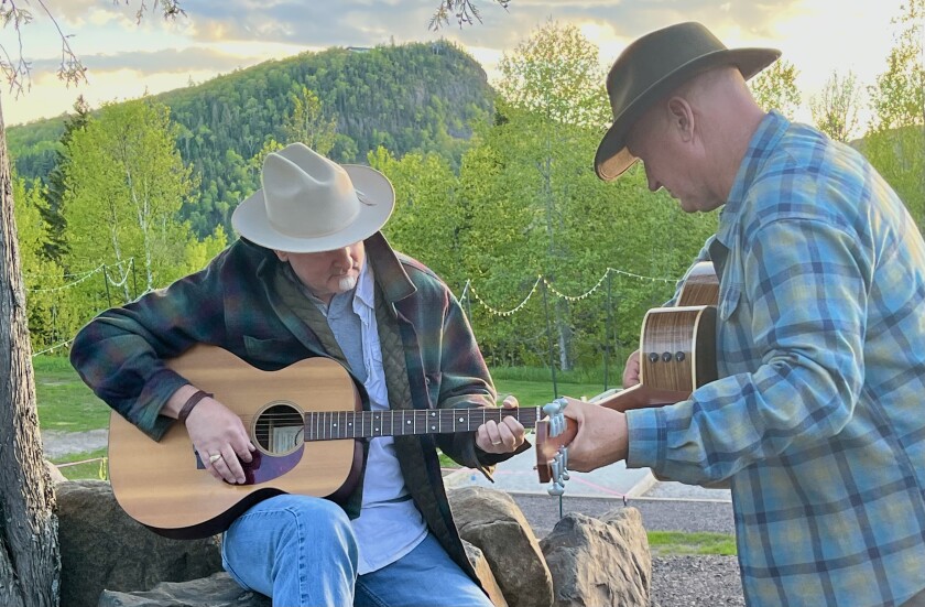 Two white-haired white men wearing coats and hats play acoustic guitars with wooded mountain visible in background.