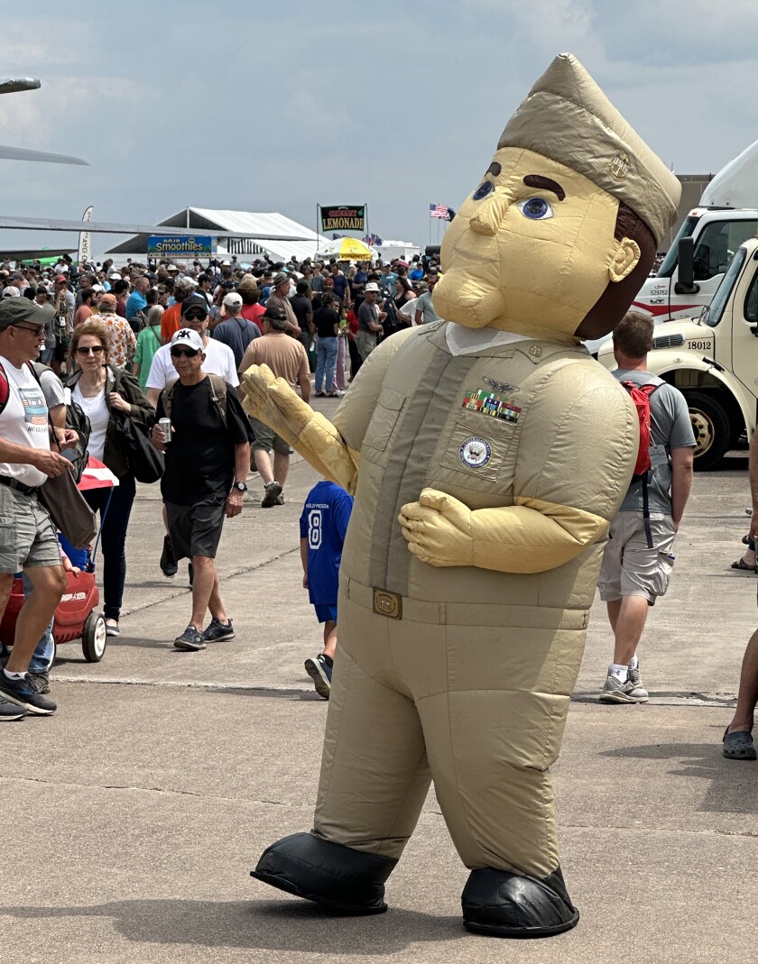 Inflatable mascot depicting Navy service member in khakis waves at a crowd of passerby on a concrete surface.