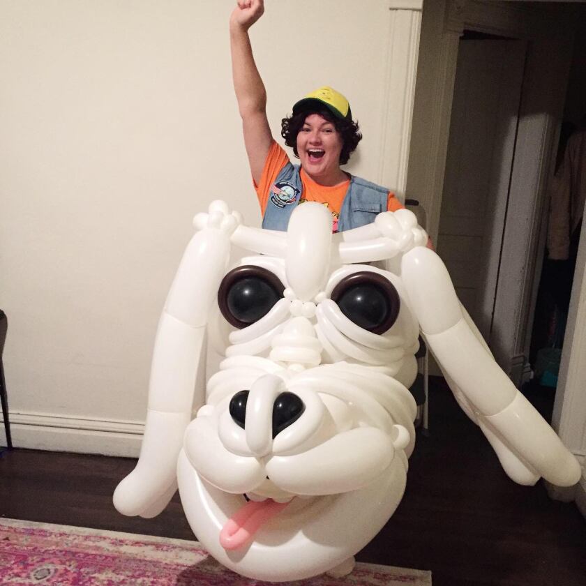 Laural Schultze, a white woman dressed as a boy in denim vest and baseball cap, raises her fist in the air and smiles as she appears to ride a white dragon head made of balloons.