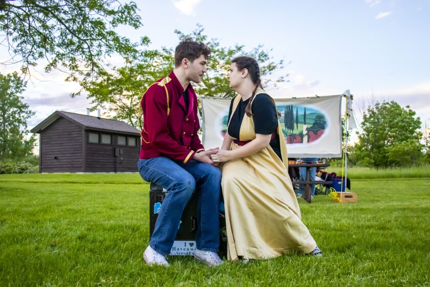 A white man and woman in colorful costume sit together on a suitcase in a grassy open-air setting with a brown wooden building, as well as a canvas painted with hilly exterior scene, visible in the background. They clasp hands and look into one another's eyes.