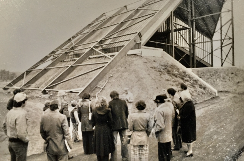 Small crowd of people stand listening to a man speak in front of a construction site featuring a tall, slanted wall.