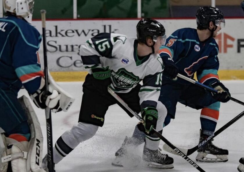Two hockey players stand next two each other during a game. They are in front of the goalie and ice is spraying up in the air.
