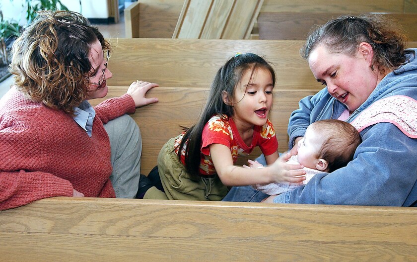 Two women tend to children, one infant and one toddlers, while sitting on a pew.