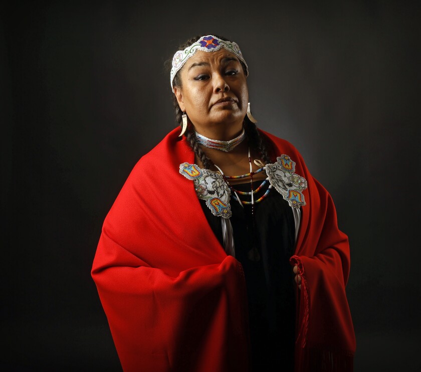 A woman wears a red blanket wrapped around her shoulder and is adorned with traditional Native American regalia.