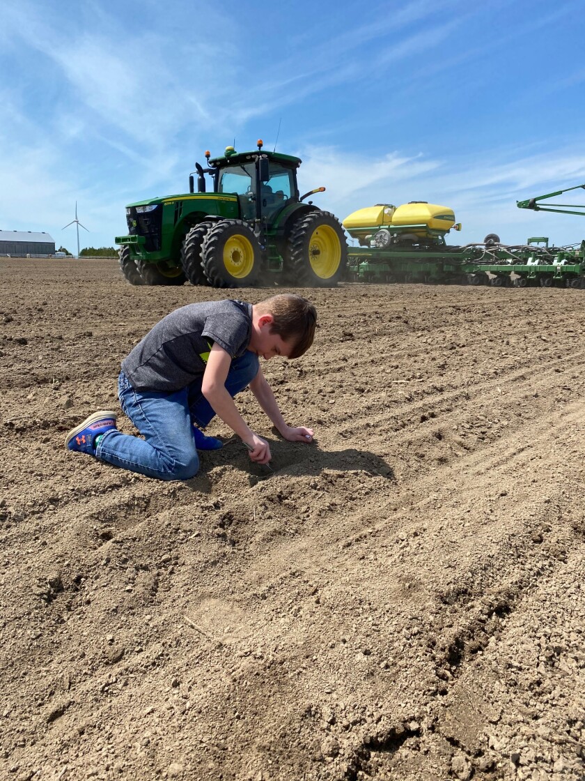 A young boy examines the soil of a furrowed field. A John Deere tractor and planter is behind him.