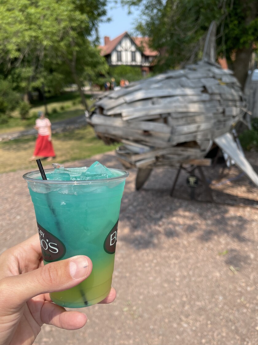 White man's hand holds a blue-and-yellow shaded cocktail in a plastic glass, with large wooden shark sculpture visible in background.