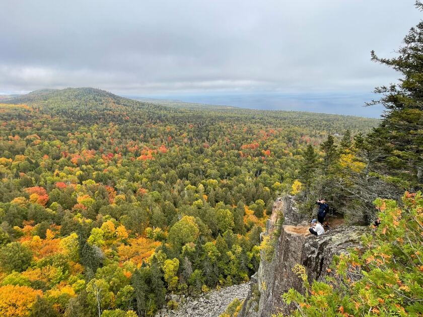 A wide expanse of fall foliage encompasses a low mountain in the background to the left. Closer to the right foreground, two hikers appear on a rocky outcrop.