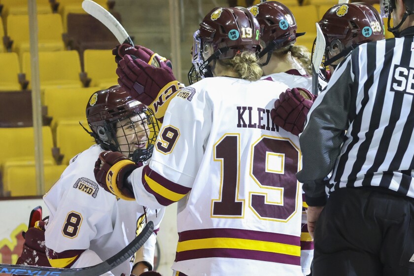 Women hockey players in purple and yellow uniforms compete against players in white and maroon uniforms on an indoor ice sheet.