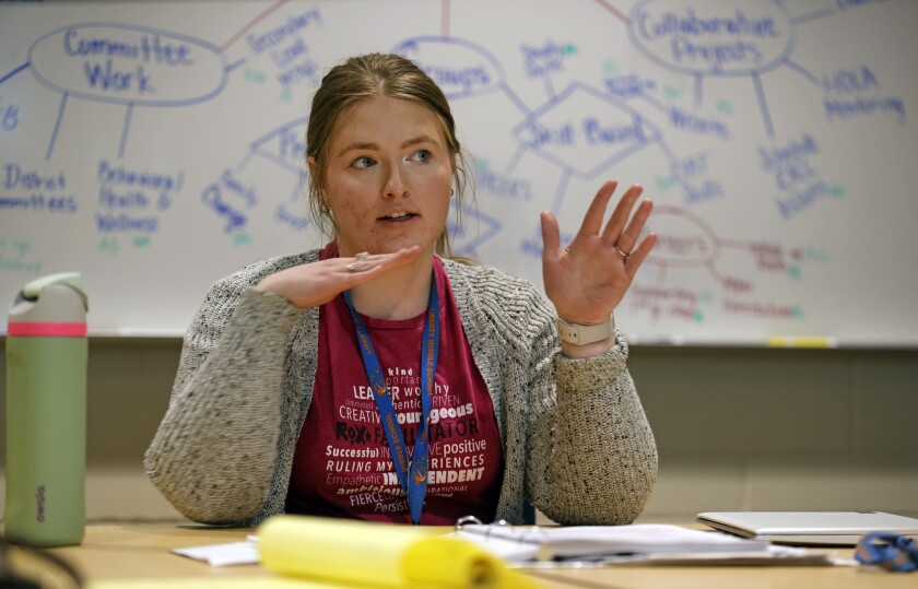 A woman in a red shirt and neutral cardigan rests her elbows on a table and gestures with both hands. She has papers in front of her and a busy whiteboard is filled with a flow chart behind her.