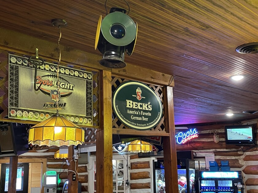Interior view of a restaurant and bar, with log walls visible in the background. In the foreground are lamps and beer signs.