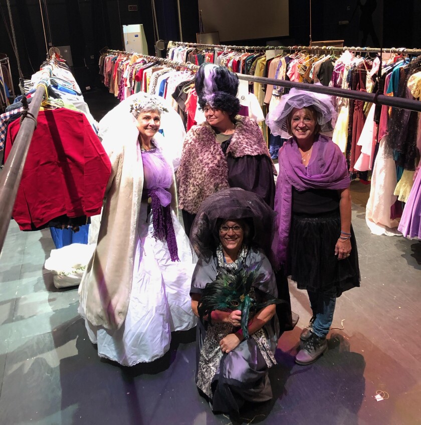 Four people pose smiling in flamboyant vintage-style dresses in front of three racks of costume pieces on a stage, seen from side stage.