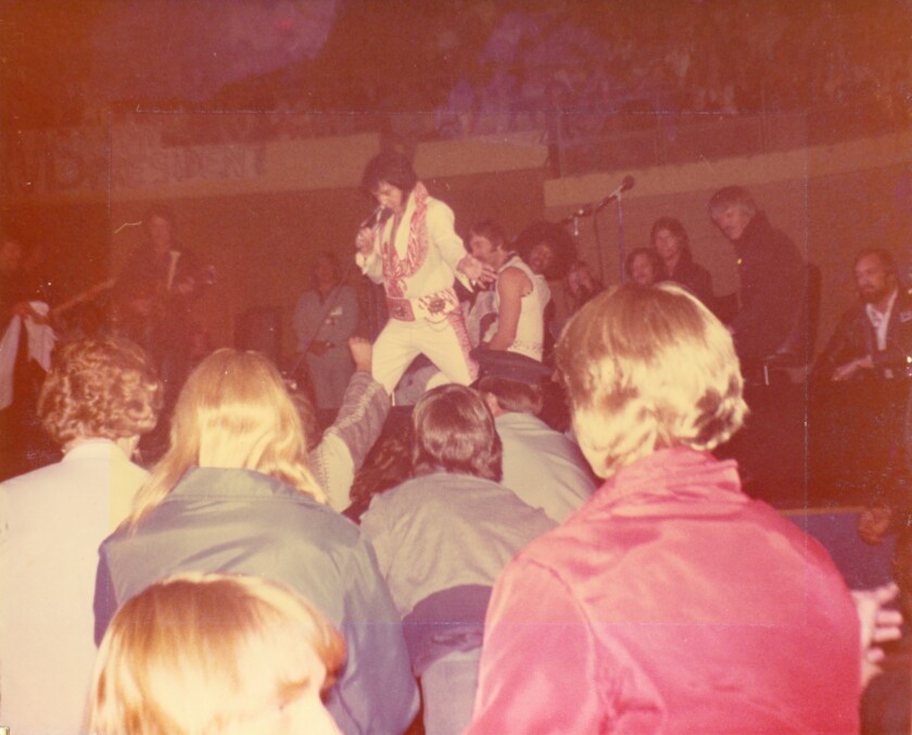 Amateur photo shows Elvis Presley, in white jumpsuit, performing onstage with several concertgoers' heads visible in the foreground.