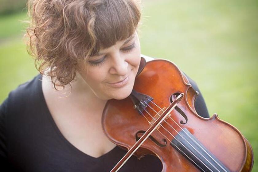 Woman with brown curly hair and black v-neck blouse playing violin in front of out-of-focus green outdoor background.
