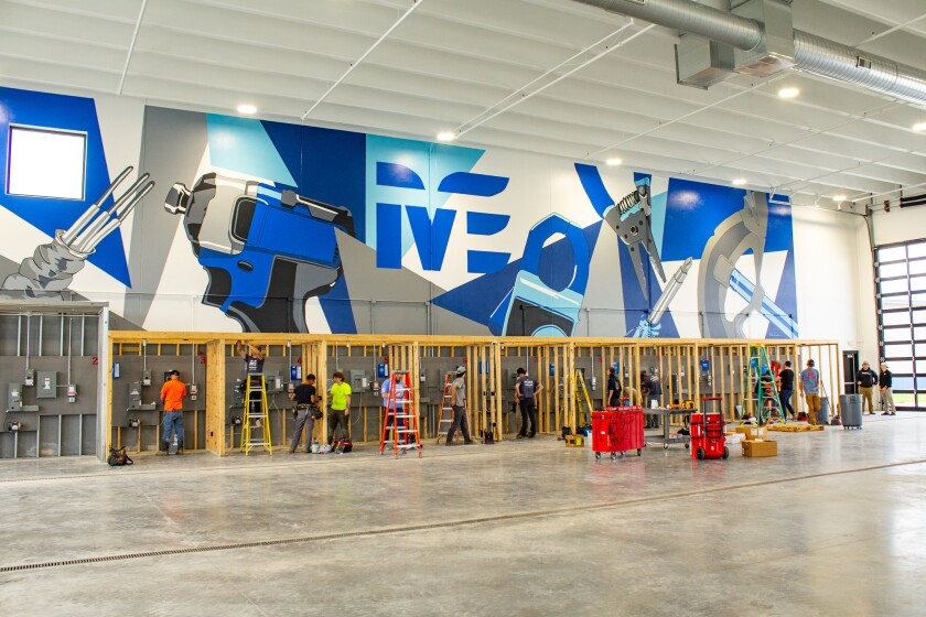 Students work at wiring lab stations, set up in a cavernous building with concrete floors and a blue, white and gray mural on the wall.
