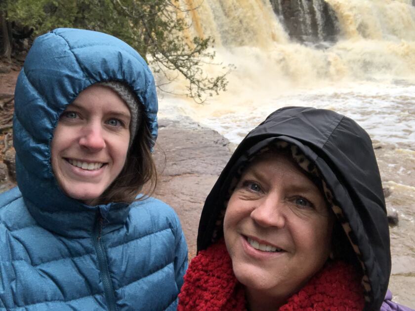 A young white woman and an older white woman stand together, smiling in hooded jackets, in front of a waterfall.