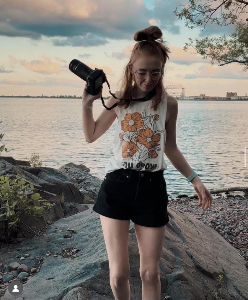 Woman wearing sleeveless floral tee holds camera standing on rocky beach with Aerial Lift Bridge visible in background.