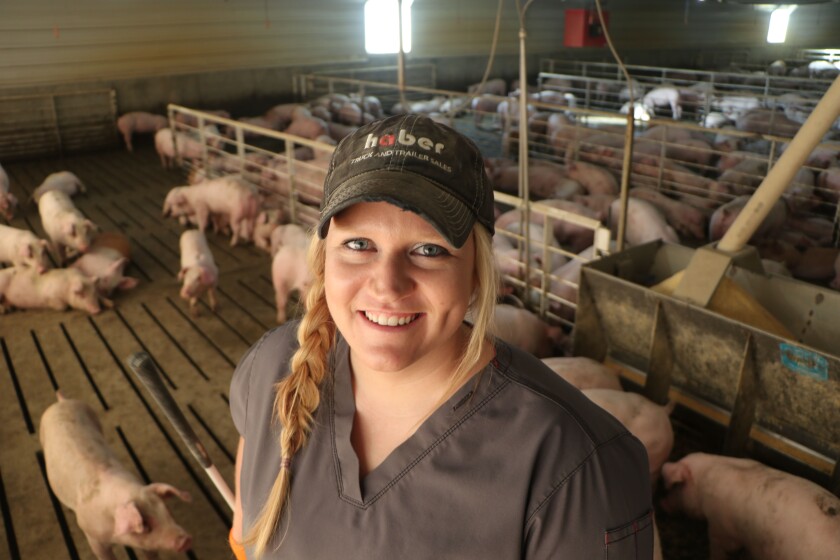 A smiling young blonde woman is flanked by pigs in pens she tends to daily in TSL, Inc., a barn she built with her husband.