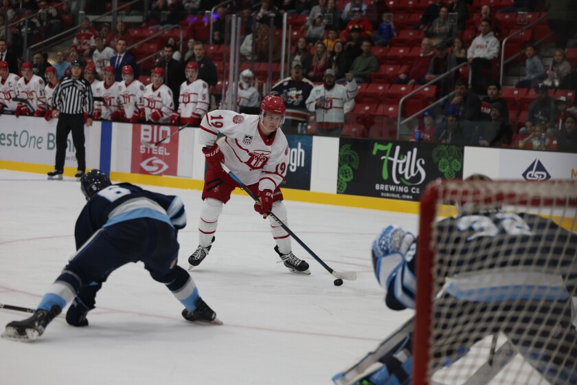 A player in a white and red hockey uniform handles the puck and prepares to shoot past a defender and a goaltender wearing white, light blue, and navy.