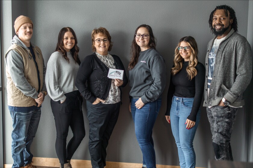 A man in a beanie and tan vest, woman in a light gray sweater, woman in a cardigan, woman in a dark gray sweater, woman in a black shirt, and man in a gray cardian stand in a row and pose for a photo.