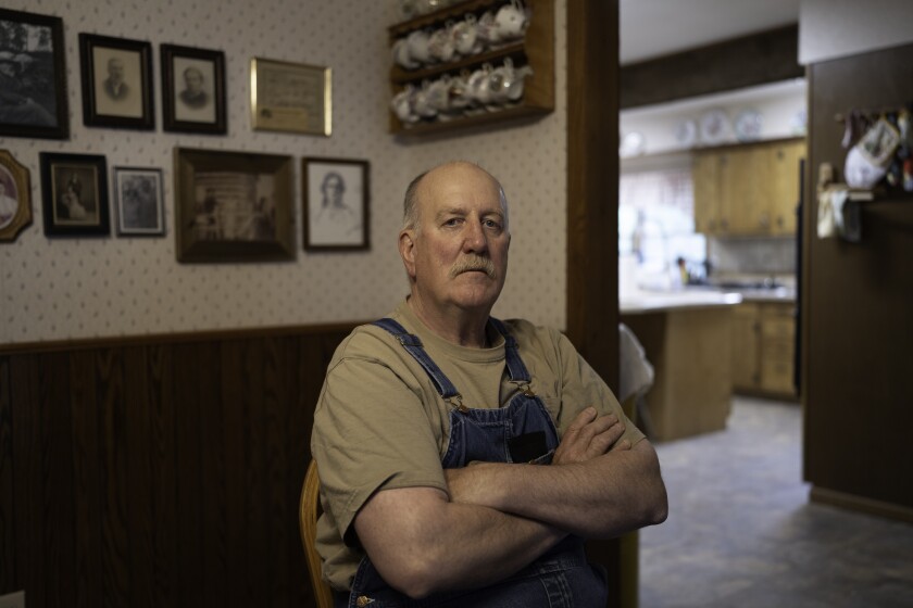 A unsmiling man in overalls sits with his arms crossed in a room with framed portraits on the wall behind him