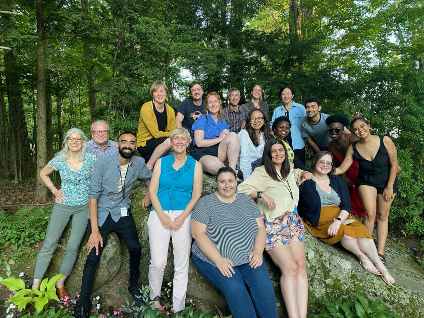 A large group of smiling people perch on a rock in a wooded setting, smiling for a portrait.