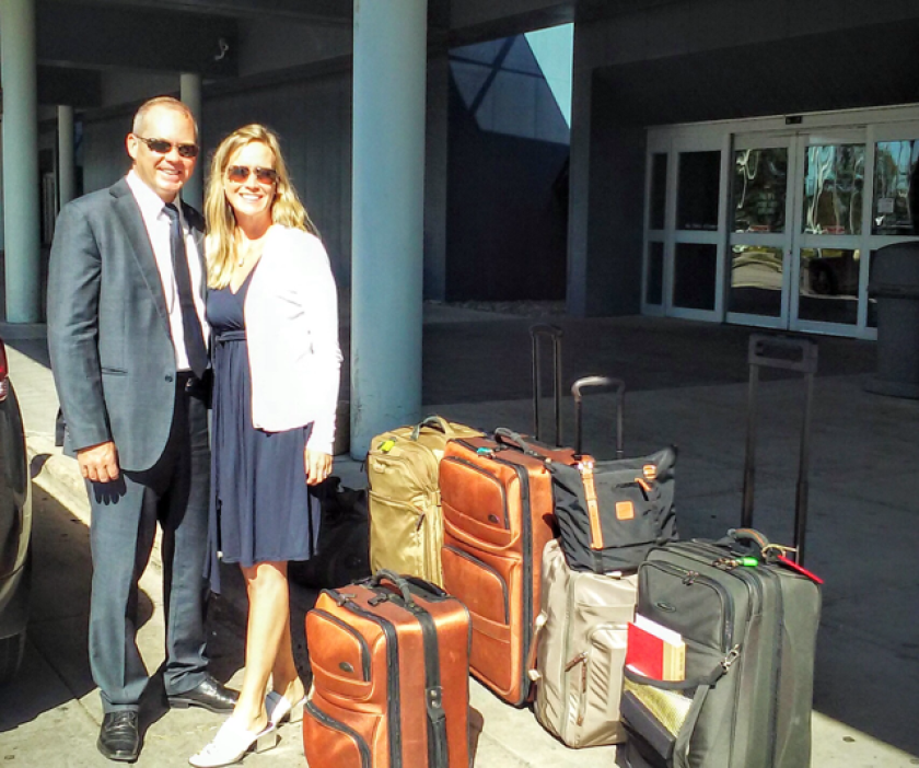 A well-dressed white couple in their 30s smile next to several large pieces of luggage.