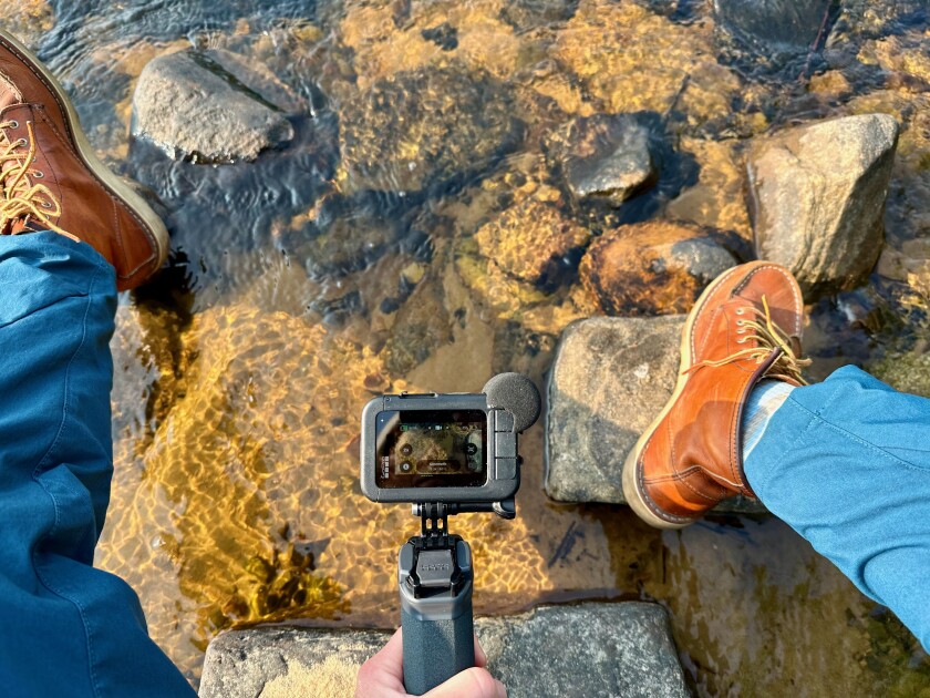 Shallow lake water is seen between a woman's legs clad in jeans and boots. A light-skinned hand holds a GoPro camera focused on the rocks visible through the water.
