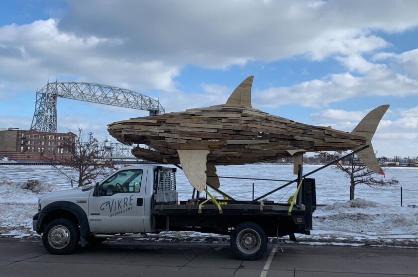 Large wooden shark sculpture sits on top of flatbed truck branded Vikre Distillery, parked in front of harbor with lift bridge in background.