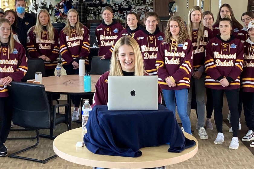 hockey player sits in front of a computer for an interview as teammates gather behind her