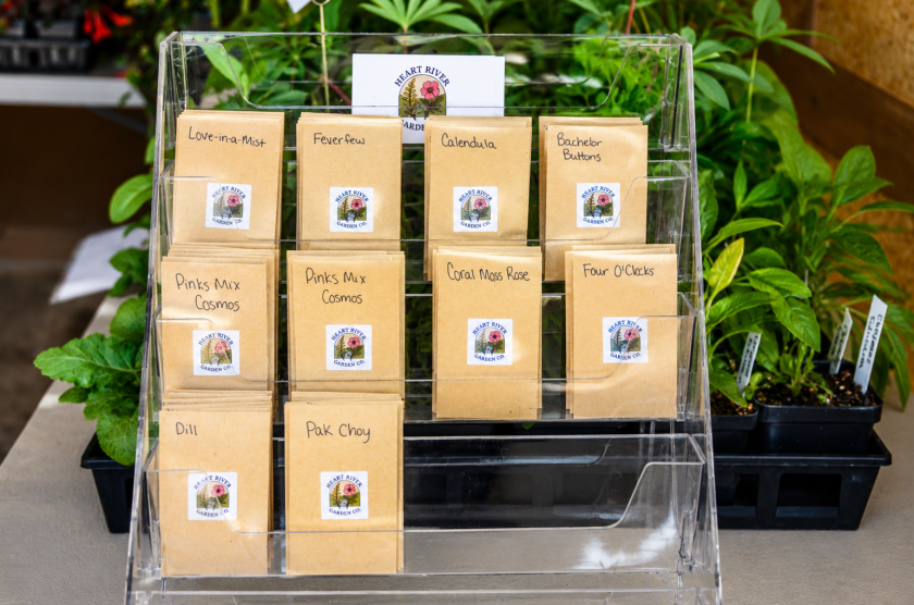 A display rack holds labeled seed packets at Heart River Garden Co near Carson, surrounded by plants inside the greenhouse.