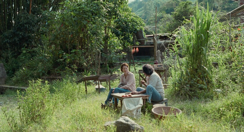 En un entorno de selva tropical verde, una mujer y un hombre se sientan juntos a hablar cerca de una mesa baja donde el hombre está trepando peces. Varias herramientas y estructuras son visibles dispersas por toda la escena.