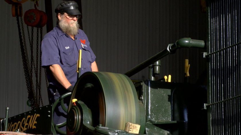A man with a gray beard and a black cap sits on the back of an old fashioned tractor.