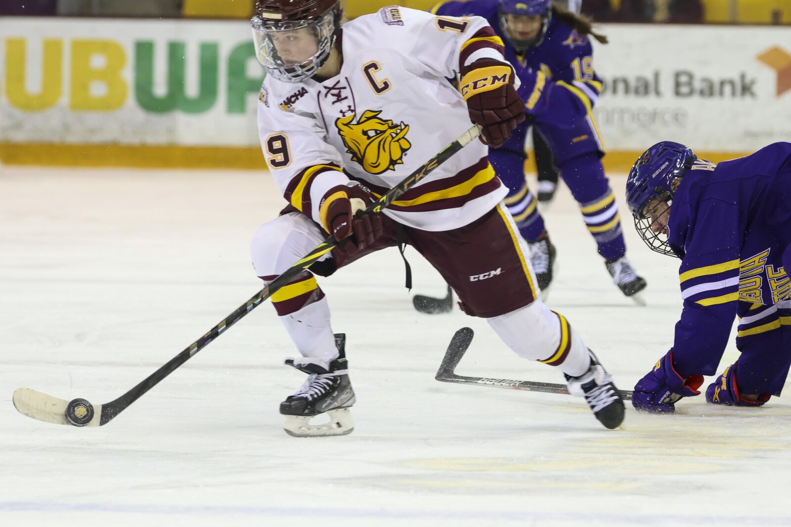 Women hockey players in purple and yellow compete against players in white and maroon at Amsoil Arena