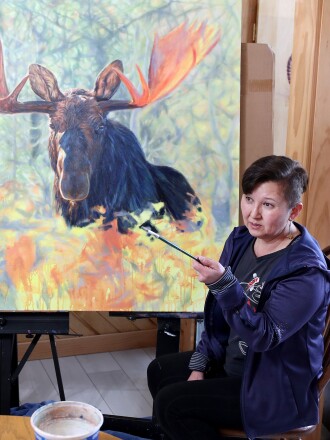 Woman holds paintbrush as she talks in front of a moose painting.