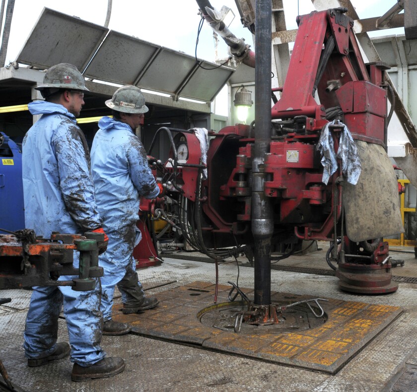 At left, two men dressed in light blue work uniforms. They are working in an industrial outdoor setting on an oil drilling rig.
