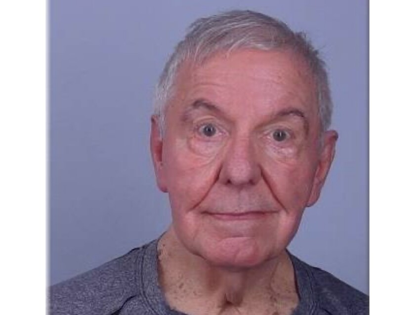 An elderly white man with eyebrows raised stares into a camera for a jail booking photo.