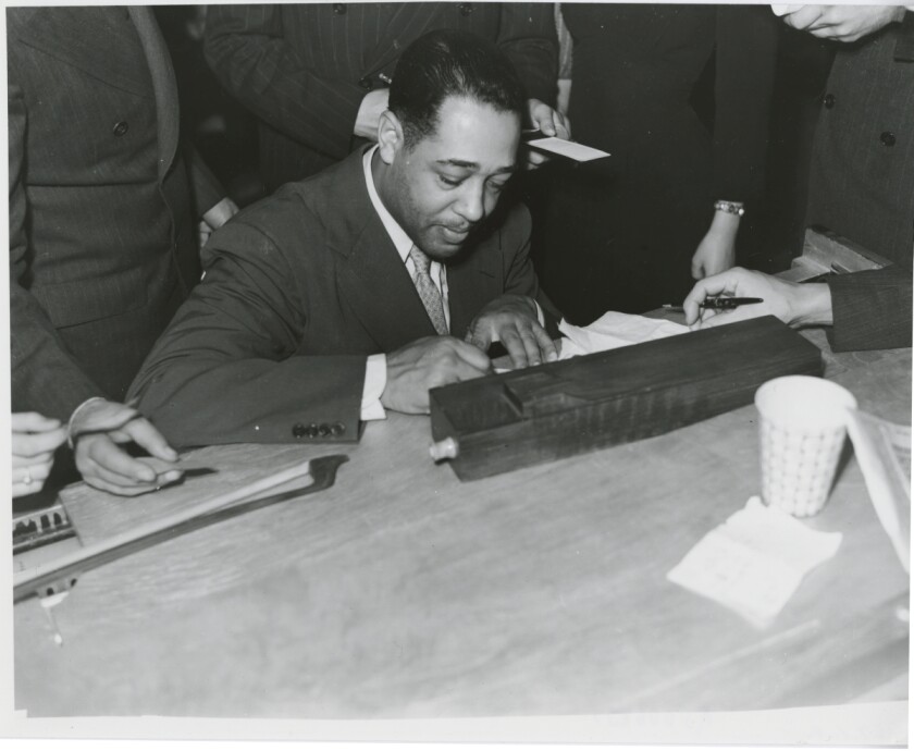 In a black and white photo, Duke Ellington signs a napkin.