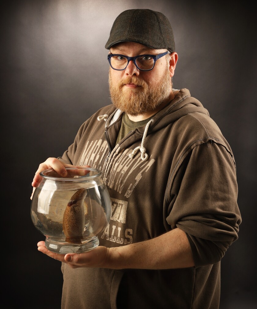 A bearded man holds a fish bowl with a round, brown amphibian inside.