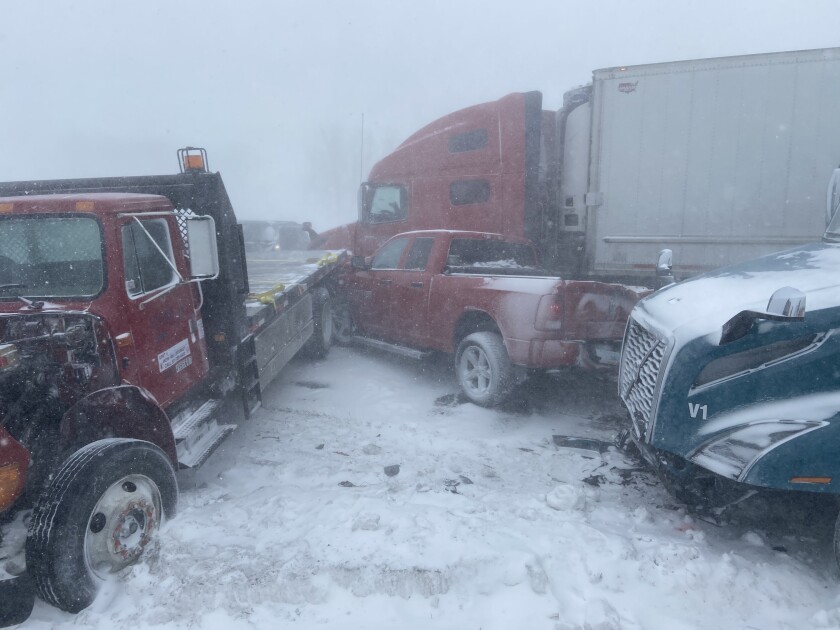 A red pickup is sandwiched between the flatbed of one semi and the tractor of another.