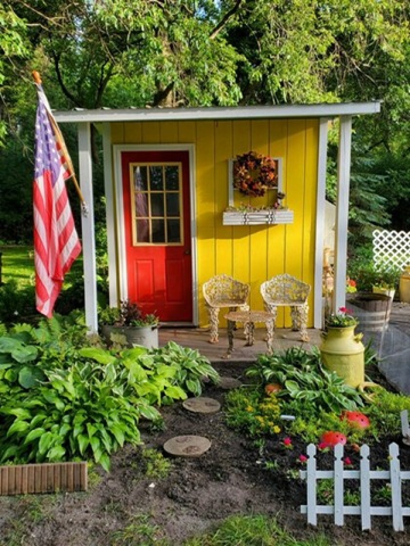 Yellow garden shed with a red door and American flag. Chairs and a table sit on a small porch in the front of the shed.