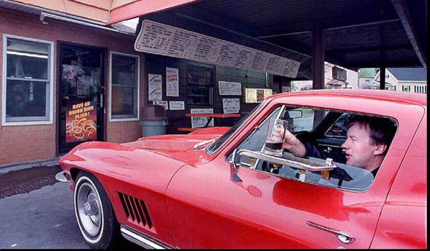 A man in a red car takes a mug of root beer from a window tray