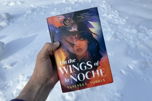 White man's hand holds book: "On the Wings of la Noche" by Vanessa L. Torres. Cover illustration features Mexican American teen with owl flying near her face.